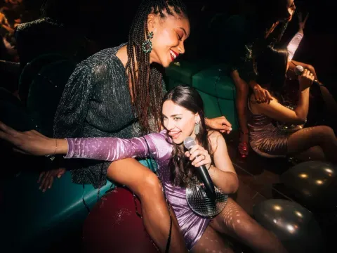 Deux jeunes femmes rient en soirée, l'une chante au micro près d'une boule disco, l'autre est assise sur une banquette turquoise
