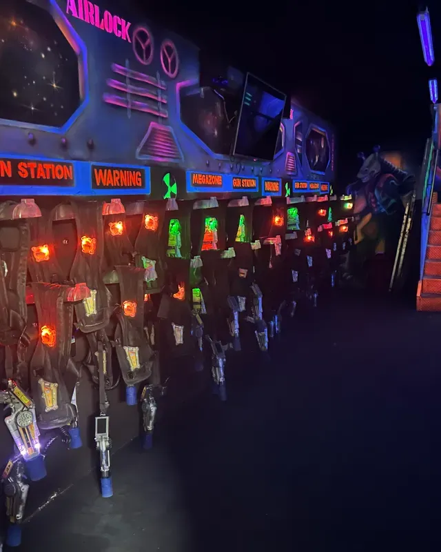 Rangée de vestes et de pistolets de laser game accrochés sous la signalétique Airlock, aux lumières néon